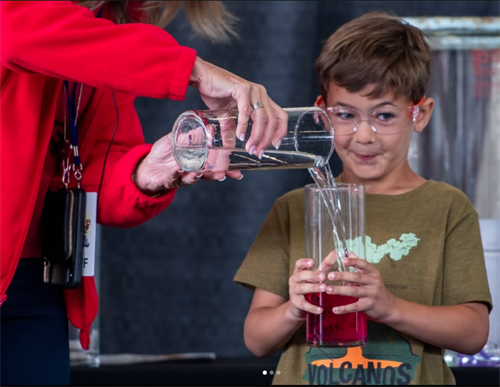 Student holding glass container while teacher pouring liquid in it.